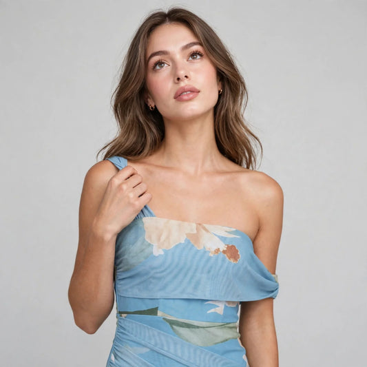 Close-up of a light blue floral one-shoulder dress with ruched bodice, hand at strap, gray studio background.