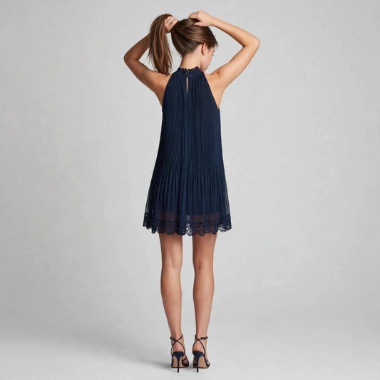 Back view of a model in a navy blue pleated halter mini dress with lace hem and strappy heels, hands in hair, gray backdrop.
