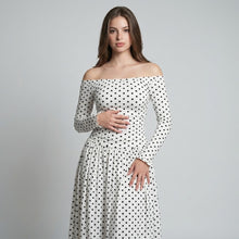 Woman in a white off-the-shoulder long-sleeve maxi dress with black polka dots and ruched bodice, gray studio background.
