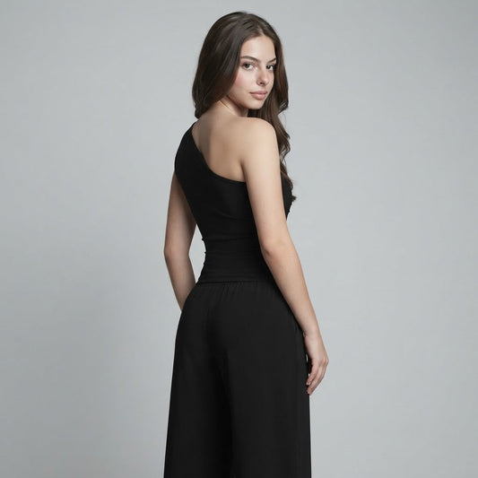 Back view of a woman looking over her shoulder in a black one-shoulder top and wide-leg pants, gray studio backdrop.