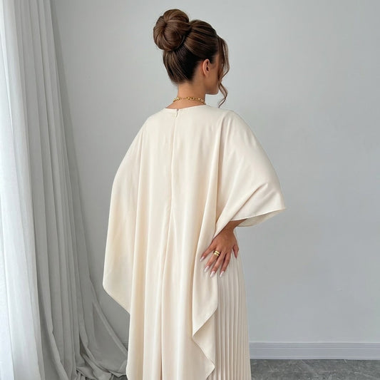Back view of a woman in an ivory cape maxi dress showing the draped cape back and pleated skirt, white curtains behind.