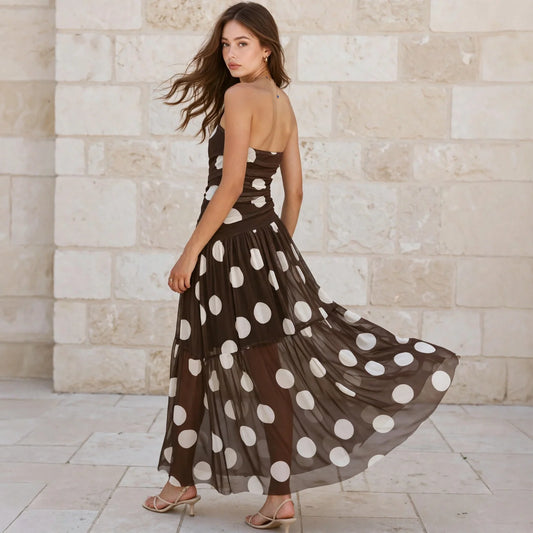 Back view of a woman walking wearing a brown strapless maxi dress with large white polka dots, beige stone wall background.