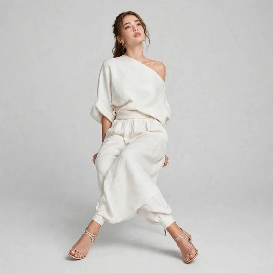 Woman posing in an ivory two-piece set with an asymmetric one-shoulder top and matching joggers, gray background.