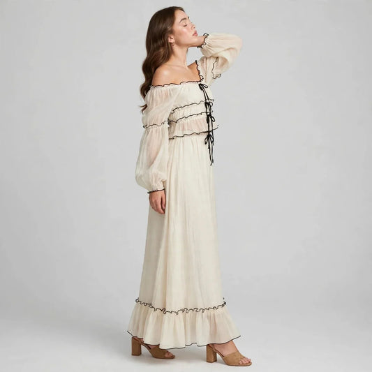 Full-length view of a woman standing in a cream off-shoulder top and long ruffled skirt set with black trim, gray studio.