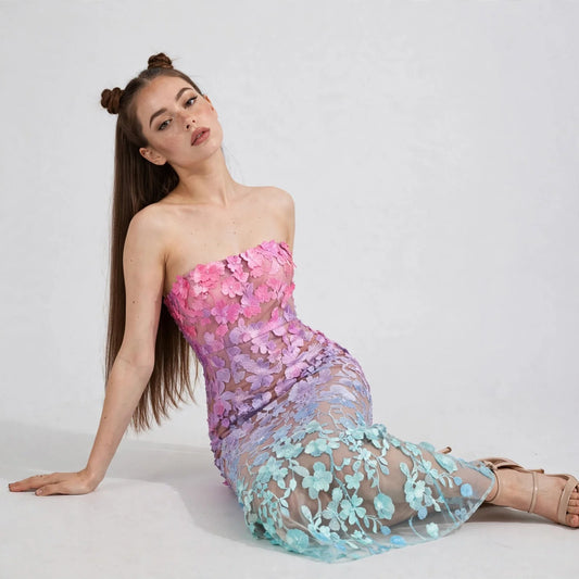 Seated woman wearing a pink-to-mint ombré floral embroidered strapless maxi dress with sheer skirt, white studio background.