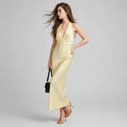 Three-quarter view of woman in pale yellow satin halter-neck maxi dress, black handbag, hair blowing, grey studio backdrop.