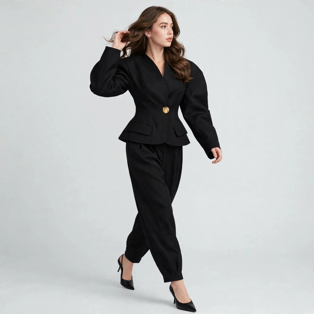 Woman walking in black puff-sleeve blazer with gold button and trousers, hand in hair, black heels, gray background.