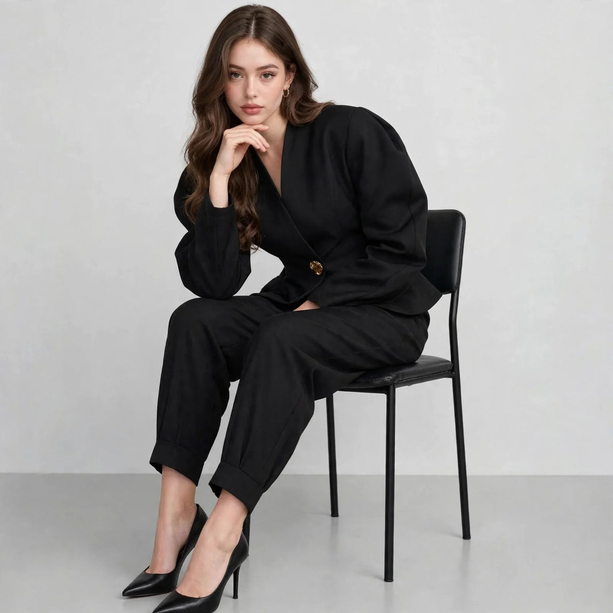 Woman sitting on a black chair wearing a black puff-sleeve blazer and trousers with pointed heels, gray studio background.