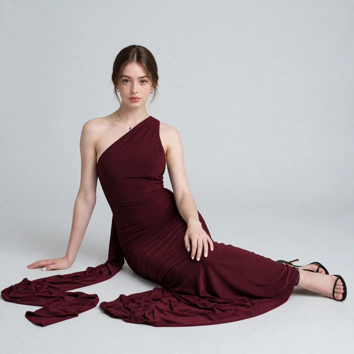 Woman seated on the floor in a burgundy one-shoulder maxi dress, skirt pooling around her, black heels, gray background.