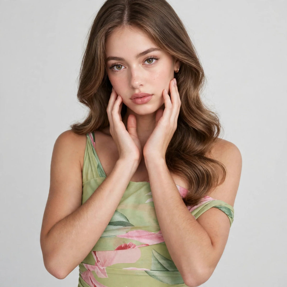 Close-up of a woman in a light green floral one-shoulder dress, hands framing her face, gray studio background.