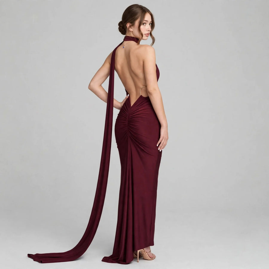 Woman modeling a burgundy one-shoulder maxi dress from the back, showing the open back and draped train, gray background.