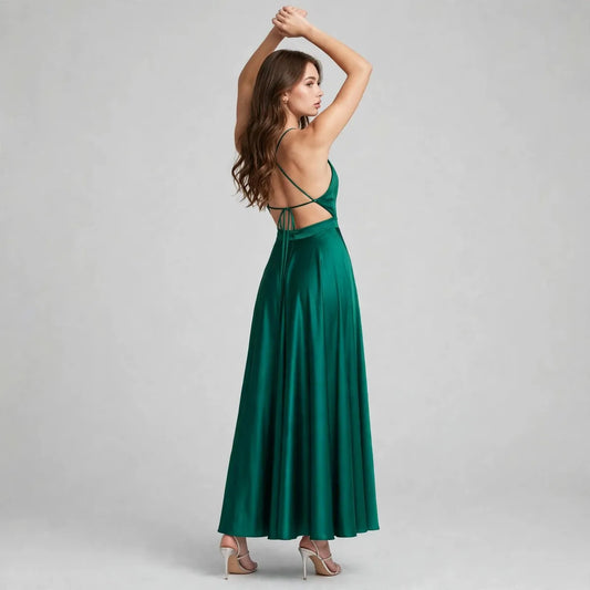 Back view of an emerald green satin maxi dress with back cutout and crisscross straps, arms raised, gray studio background.