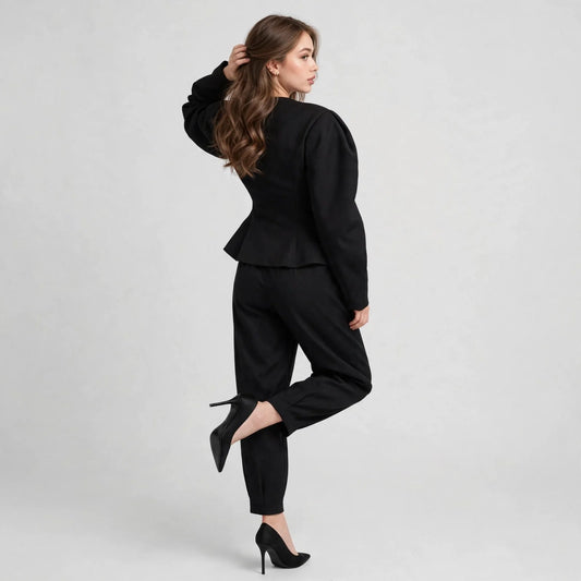 Back view of woman in black puff-sleeve peplum blazer and trousers, one leg lifted, black heels, gray background.