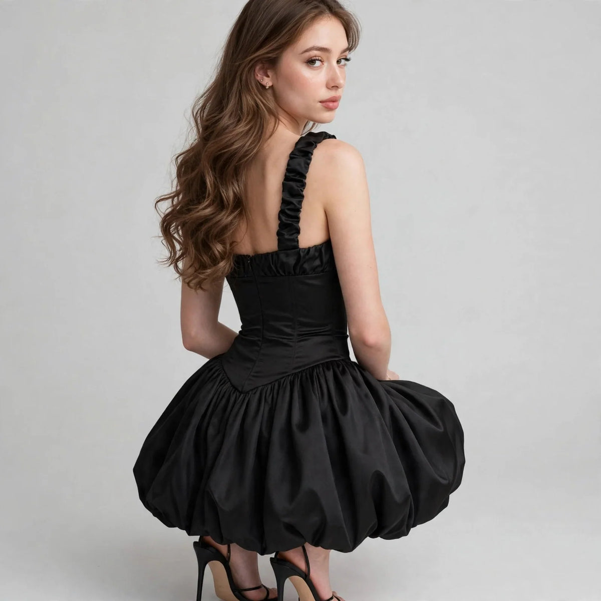Back three-quarter view of a black satin bubble hem dress with ruffled strap and corset bodice, black heels, gray studio.