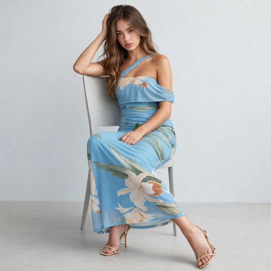 Woman sitting on a chair in a light blue floral one-shoulder maxi dress with ruched bodice, beige heels, gray background.