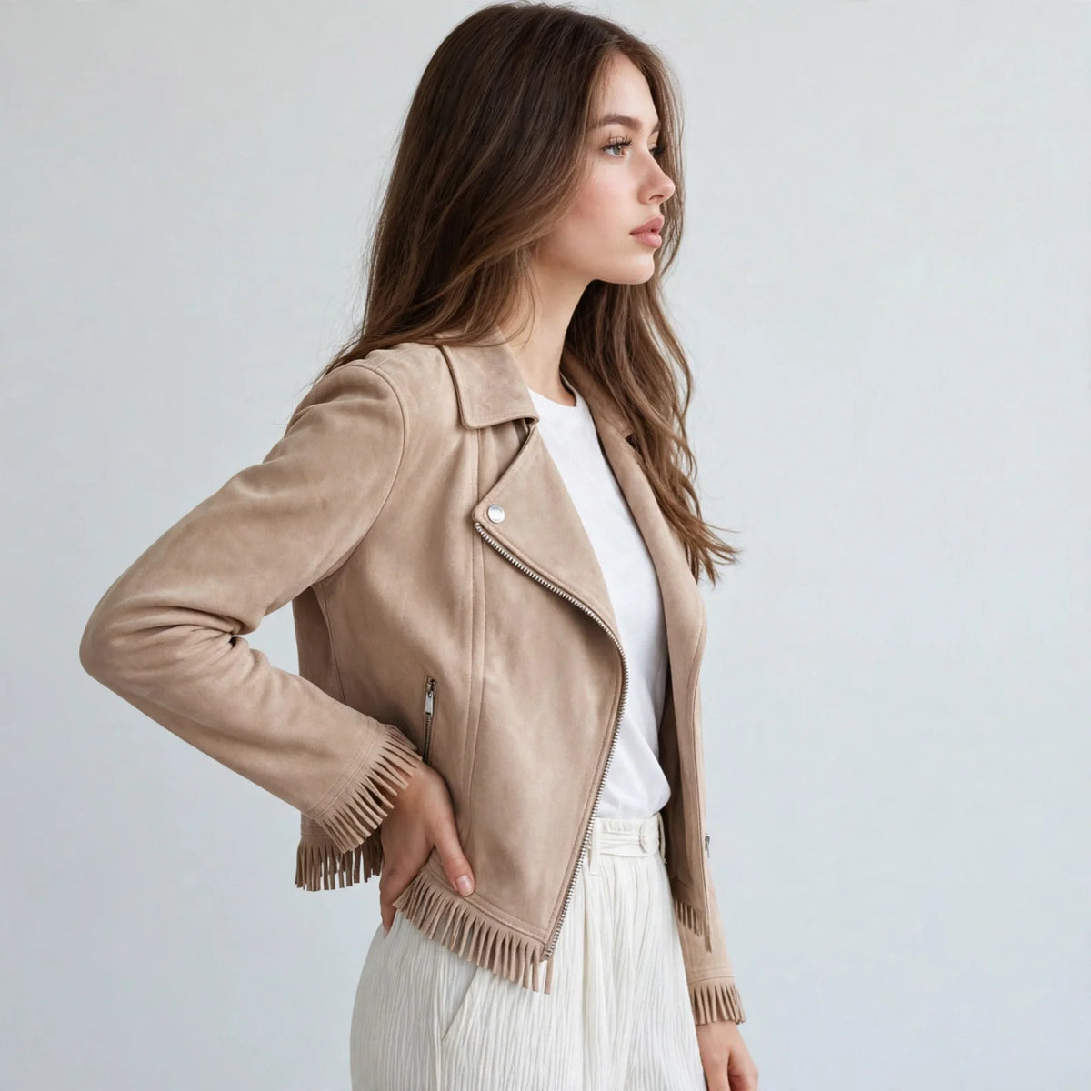 Side view of a woman with hand on hip in a beige faux suede fringe moto jacket with white top and jeans, gray studio.