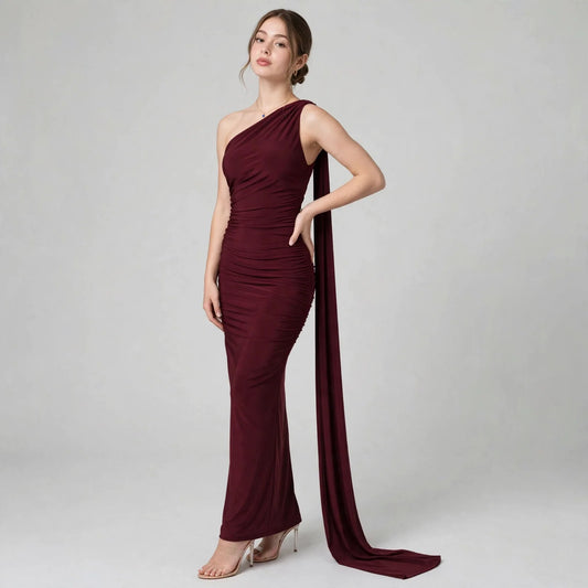 Woman modeling a floor-length burgundy one-shoulder gown with ruched detailing and a draped train, gray studio background.