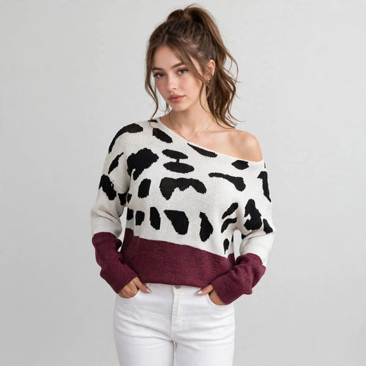 Woman in a white one-shoulder sweater with black spots and burgundy hem band, white pants, front view, gray background.