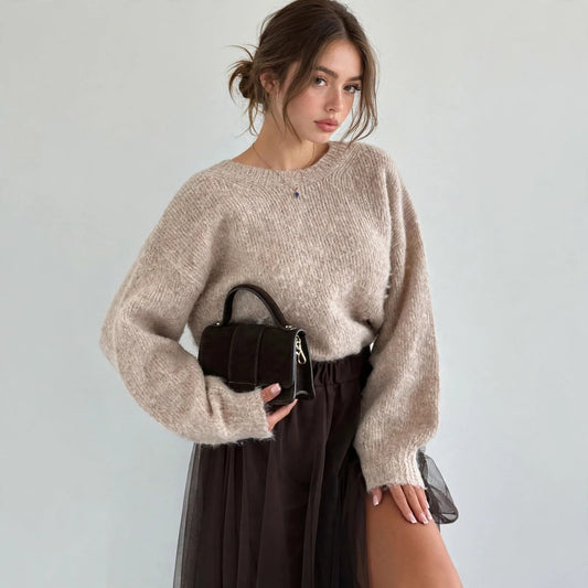 Model in beige fuzzy dropped-shoulder knit sweater holding small black handbag, three-quarter front view, studio backdrop.
