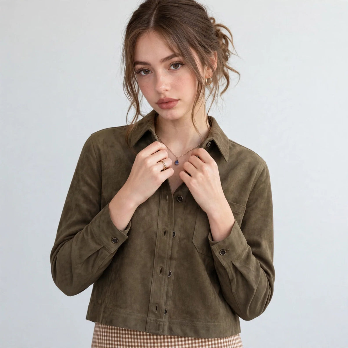 Close-up of a woman buttoning an olive cropped faux suede button-down jacket over a white top, gray background.