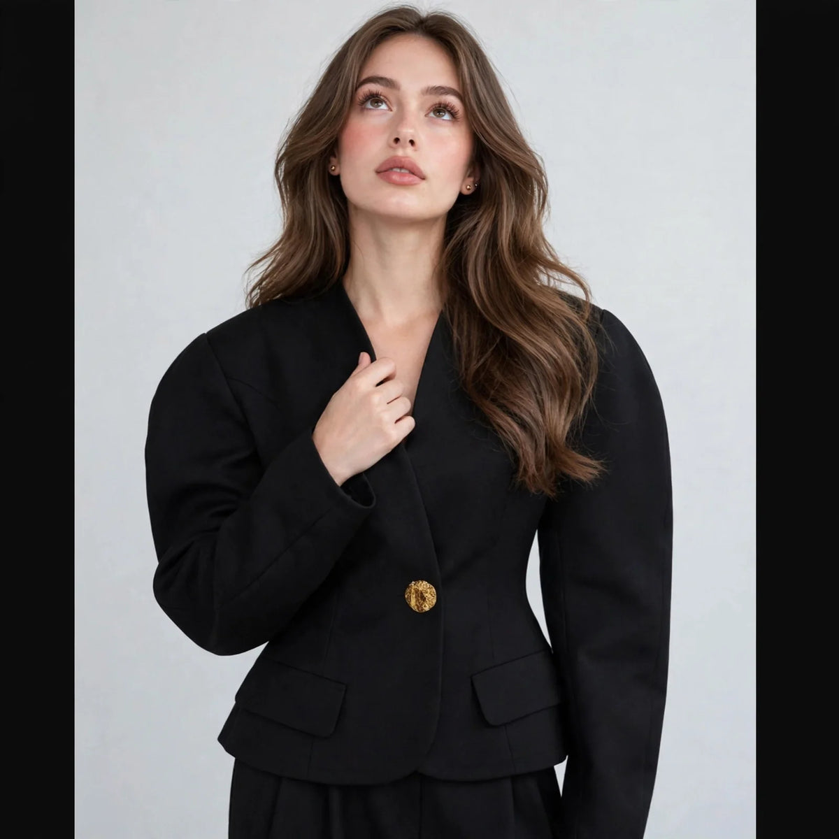 Close-up of a black tailored blazer with puff sleeves and a single gold button, worn by a woman, gray studio background.