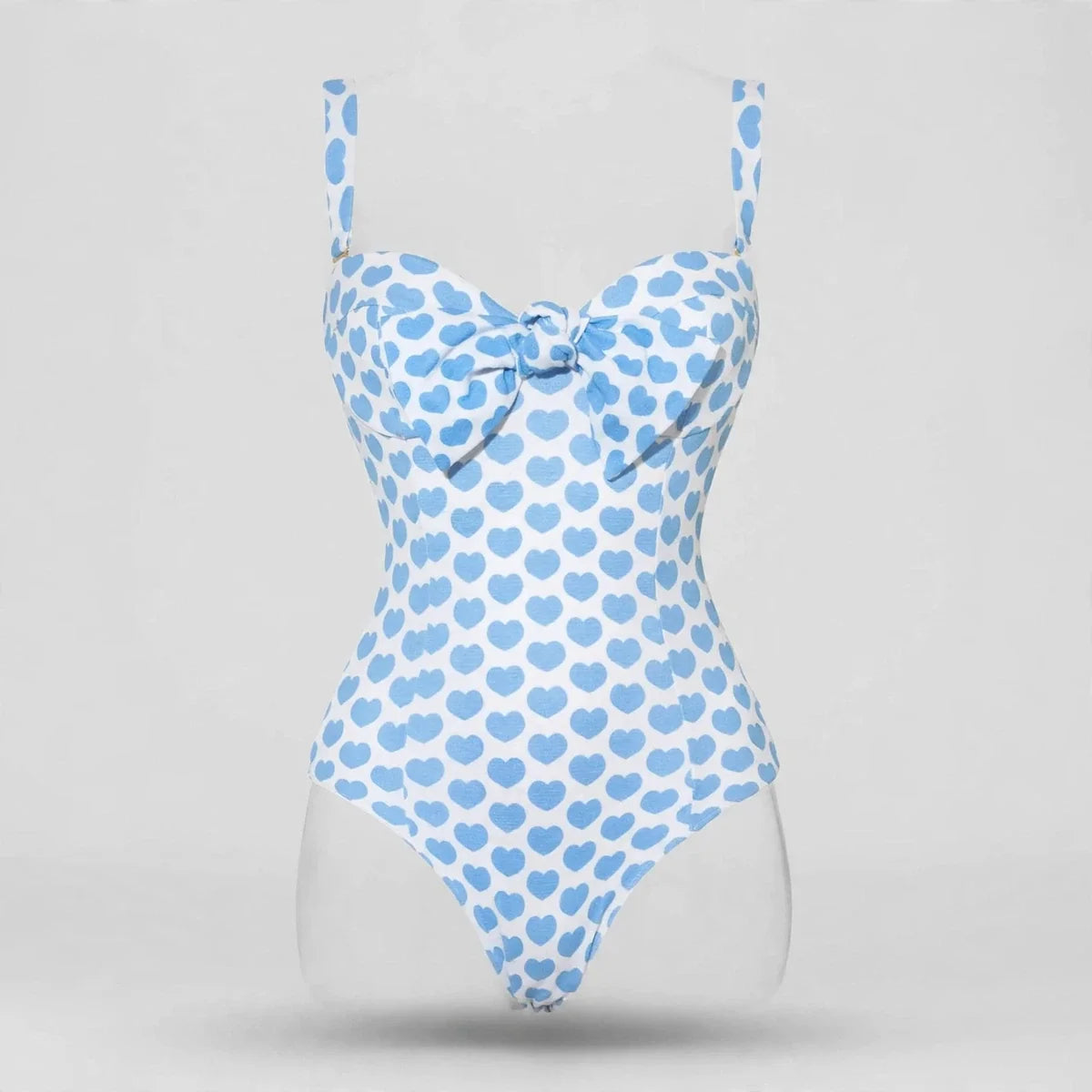 Flat lay of a blue heart-print one-piece swimsuit with sweetheart neckline and bow detail at the bust, gray background.