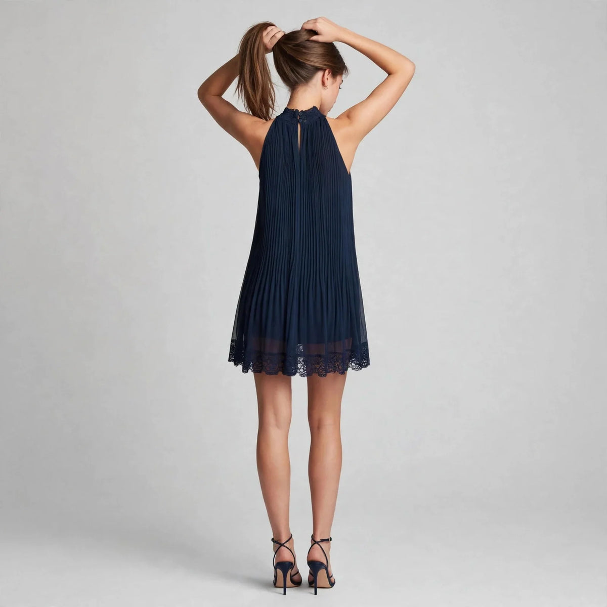 Back view of a model in a navy blue pleated halter mini dress with lace hem and strappy heels, hands in hair, gray backdrop.