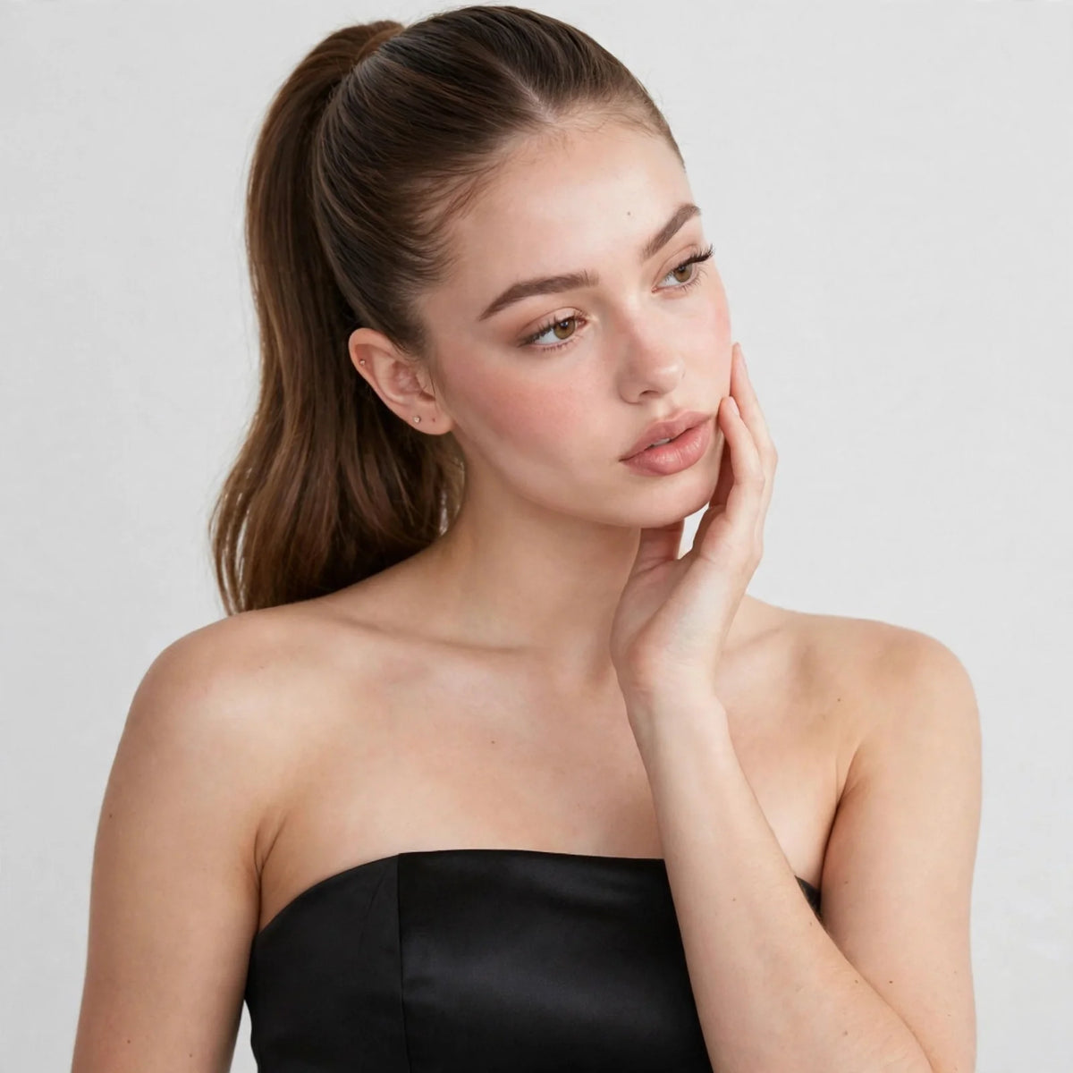 Close-up of a woman with a ponytail wearing a black strapless satin top, hand near face, gray studio background.