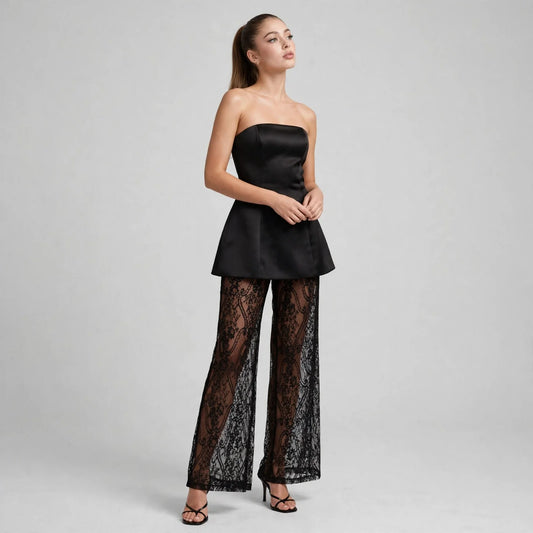 Front view of model with hands clasped in black strapless peplum tube top and sheer black lace wide-leg pants, grey backdrop