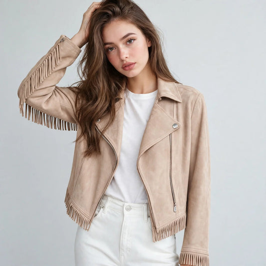 Woman with hand in hair wearing a beige faux suede fringe moto jacket over a white top and white jeans, gray studio backdrop.