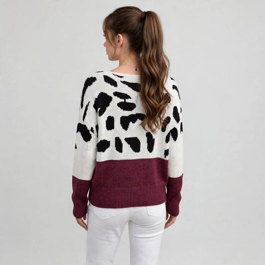 Back view of a woman in a white one-shoulder sweater with black spots and burgundy hem band, white pants, gray background.