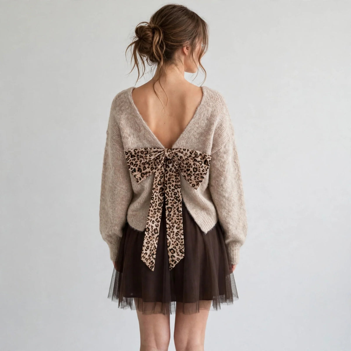Back view of model in beige fuzzy dropped-shoulder sweater with leopard bow at open back neckline, full-length studio shot.