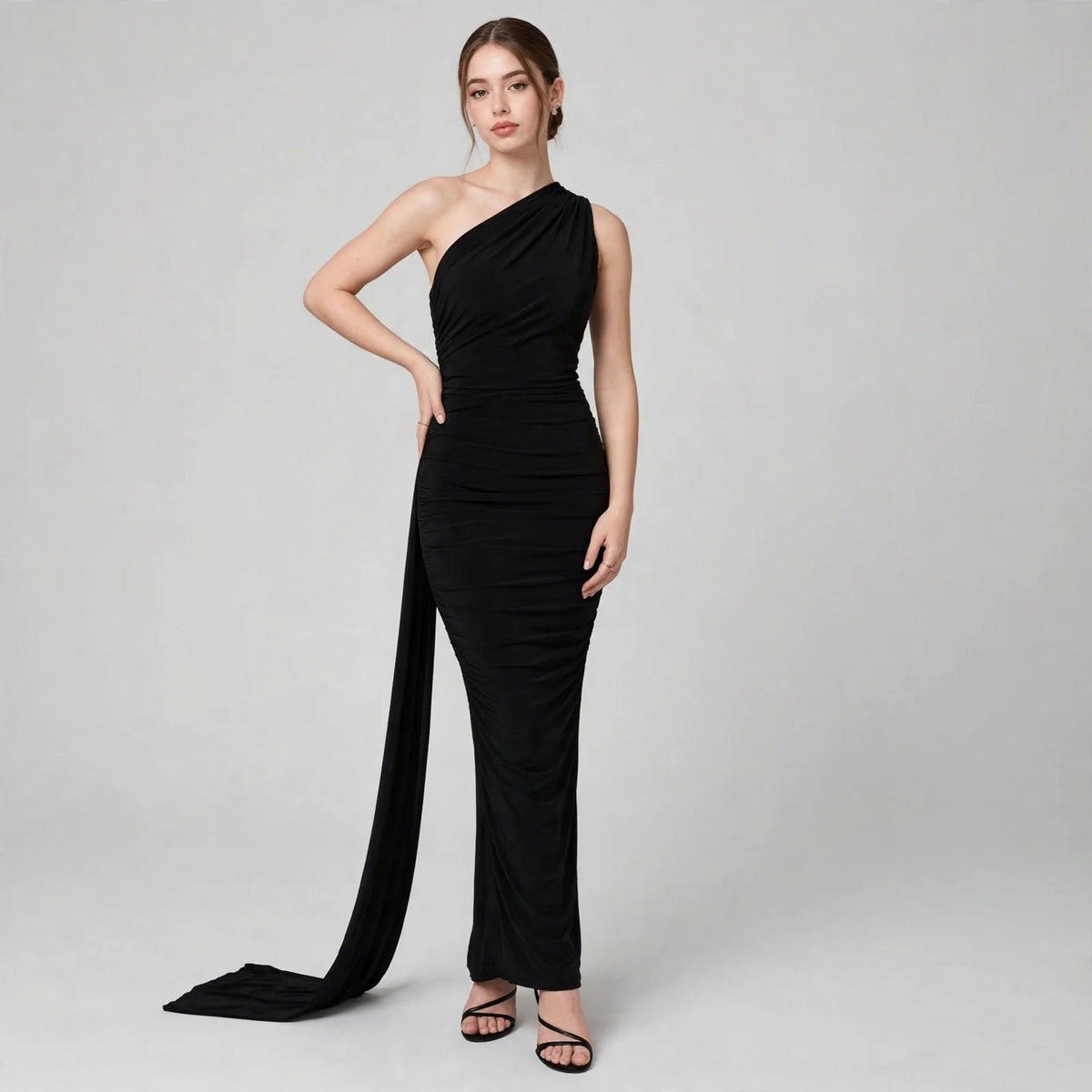 Woman posing in a black one-shoulder ruched maxi dress with a long flowing train and black heels, gray studio background.