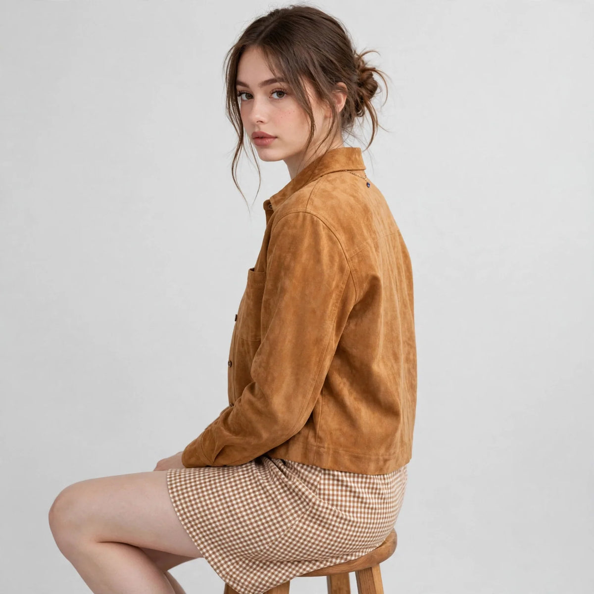 Woman seated sideways showing the back of a camel cropped faux suede jacket with a checkered skirt, gray background.