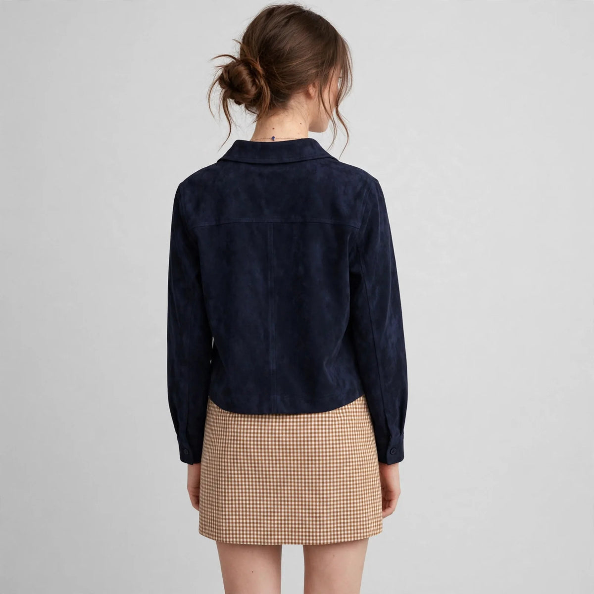 Woman viewed from behind wearing a navy cropped faux suede button-down jacket and a checkered skirt, gray background.