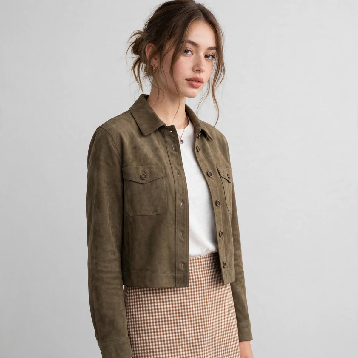 Woman standing in an olive cropped faux suede button-down jacket with a white top and checkered skirt, gray background.