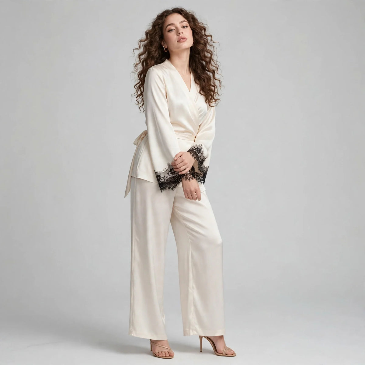 Front view of model in ivory satin wrap top with black lace cuffs and matching wide-leg pants, barefoot on grey background