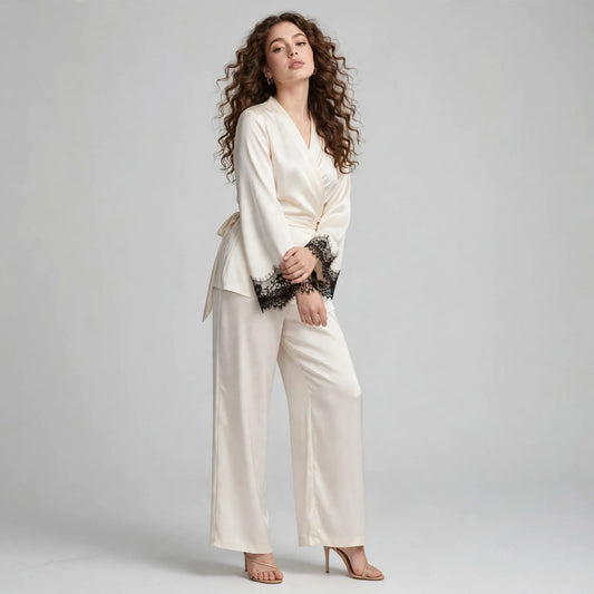 Front view of model in ivory satin wrap top with black lace cuffs and matching wide-leg pants, barefoot on grey background