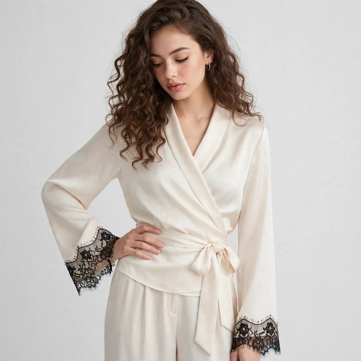 Close-up front view of model with hand on hip in ivory satin wrap top with black lace cuff trim and wide-leg pants