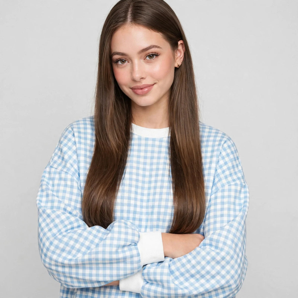 Close-up front view of a light blue gingham sweatshirt with white ribbed cuffs, model with arms crossed, gray studio.