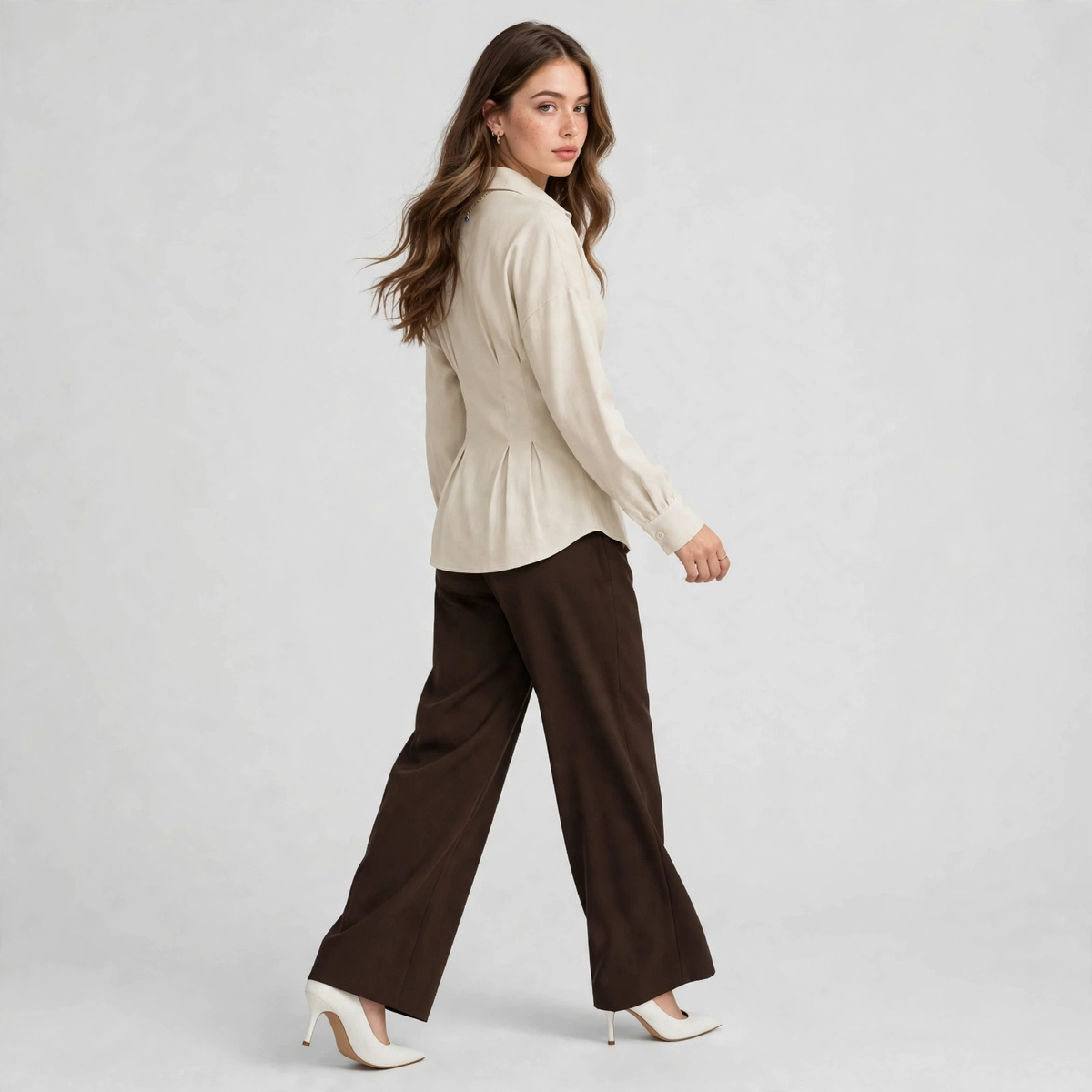 Three-quarter back view of a woman walking in a cream ruched peplum shirt and dark brown wide-leg trousers, gray studio.
