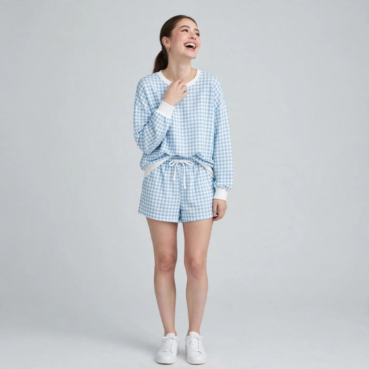 Model smiling in a blue and white gingham sweatshirt and shorts set with white trim, paired with white sneakers, studio.