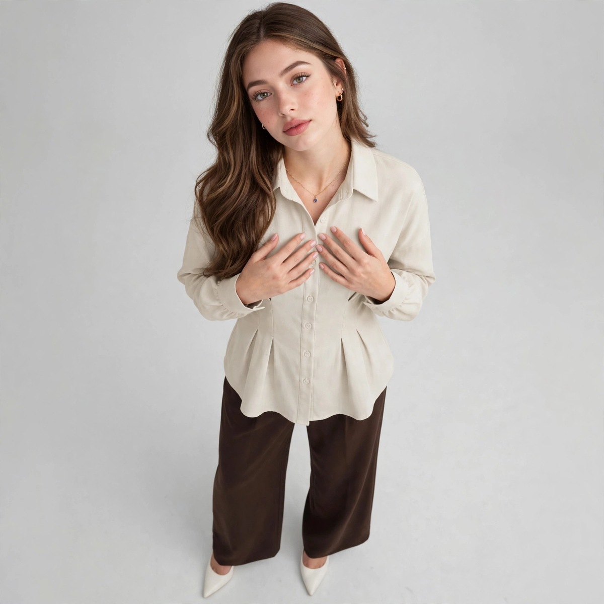 High-angle view of a woman with hands on chest wearing a cream ruched button-up peplum shirt and brown trousers, gray studio.