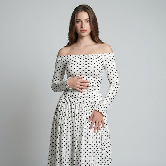 Woman in a white off-the-shoulder long-sleeve maxi dress with black polka dots and ruched bodice, gray studio background.