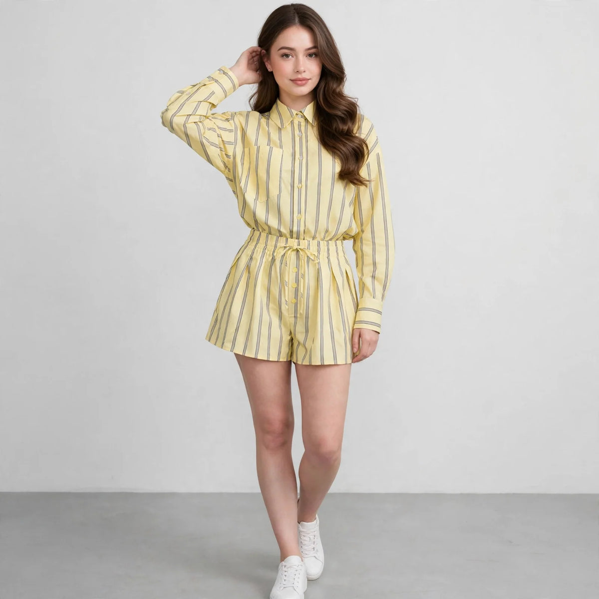 Woman in yellow and grey striped button-up shirt and shorts set, white sneakers, hand in hair, front view, light backdrop.