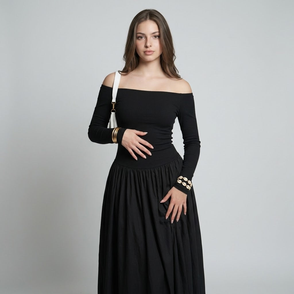 Woman in a black off-the-shoulder maxi dress with gathered waist and pleated skirt, gold bracelets, gray studio background.