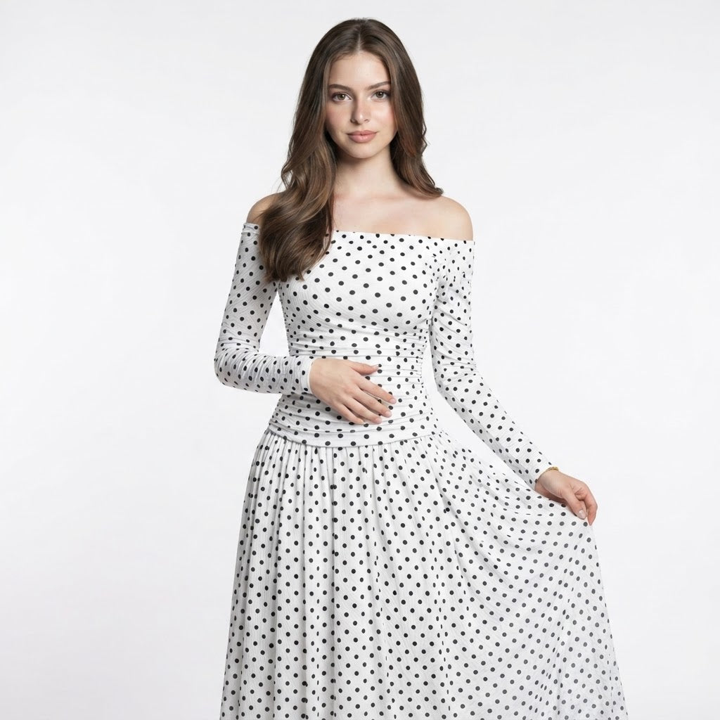 Woman in a white long-sleeve off-the-shoulder maxi dress with black polka dots and flowing skirt, white studio background.