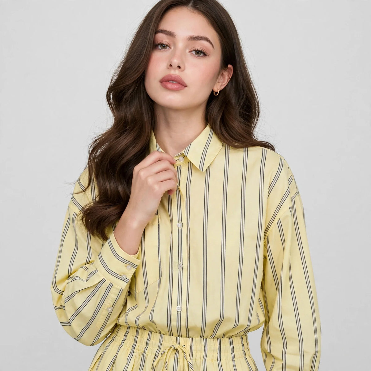 Close-up of woman in yellow and grey striped button-up shirt, collar and front buttons, hand near neck, light backdrop.