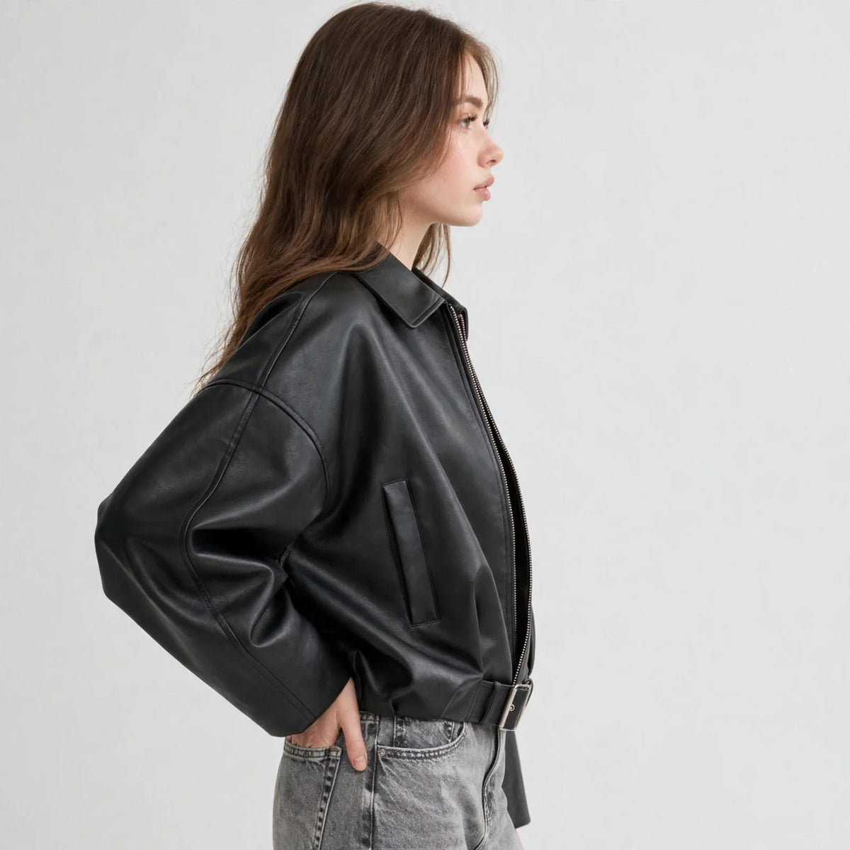 Side view of model wearing a black cropped faux leather zip-up jacket with belted hem and grey jeans, grey studio background