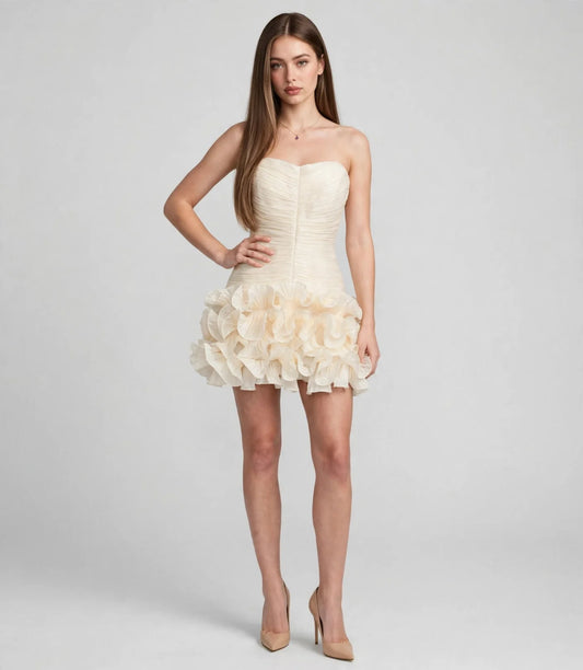 Full-body front view of a woman in a cream strapless mini dress with a ruched bodice and layered ruffle skirt, nude heels.